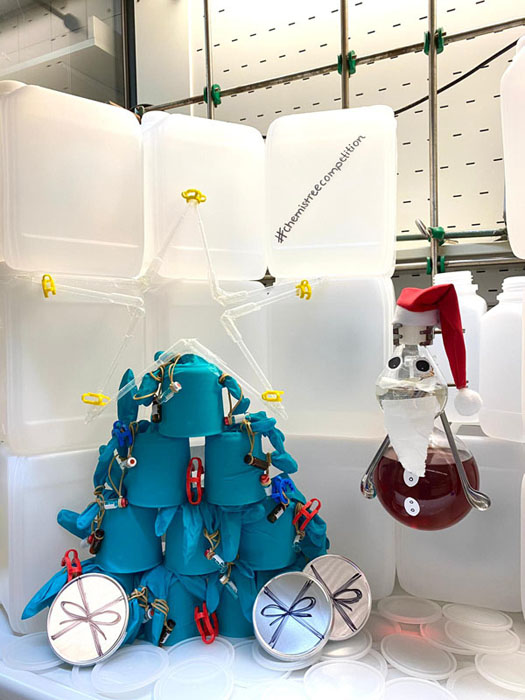 Vor einer Kulisse aus Kanistern sieht man einen ChemisTree aus grünen Labor-Handschuhen, Glasfläschchen-Schmuck, einem Stern aus Pipetten und Petrischalen-Geschenken. Und: ein Kolben-Weihnachtsmann inklusive Mütze und Rauschebart. Danke, Nina! Sie ist Azubine bei der BASF.